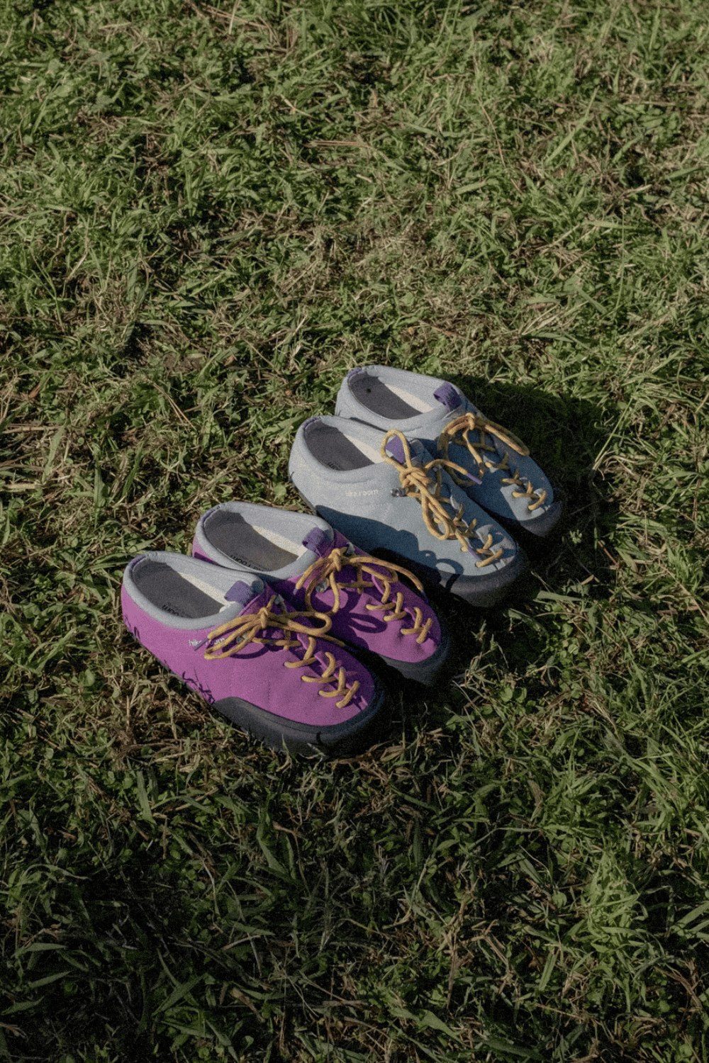 Dos pares de zapatillas destalonadas Hike Room en colores morado y gris claro con cordones amarillos descansando directamente sobre el césped verde. Descripción: Los diseños de Hike Room demuestran su enfoque funcional y estético, fusionando el equipamiento de montaña con la comodidad del calzado urbano.