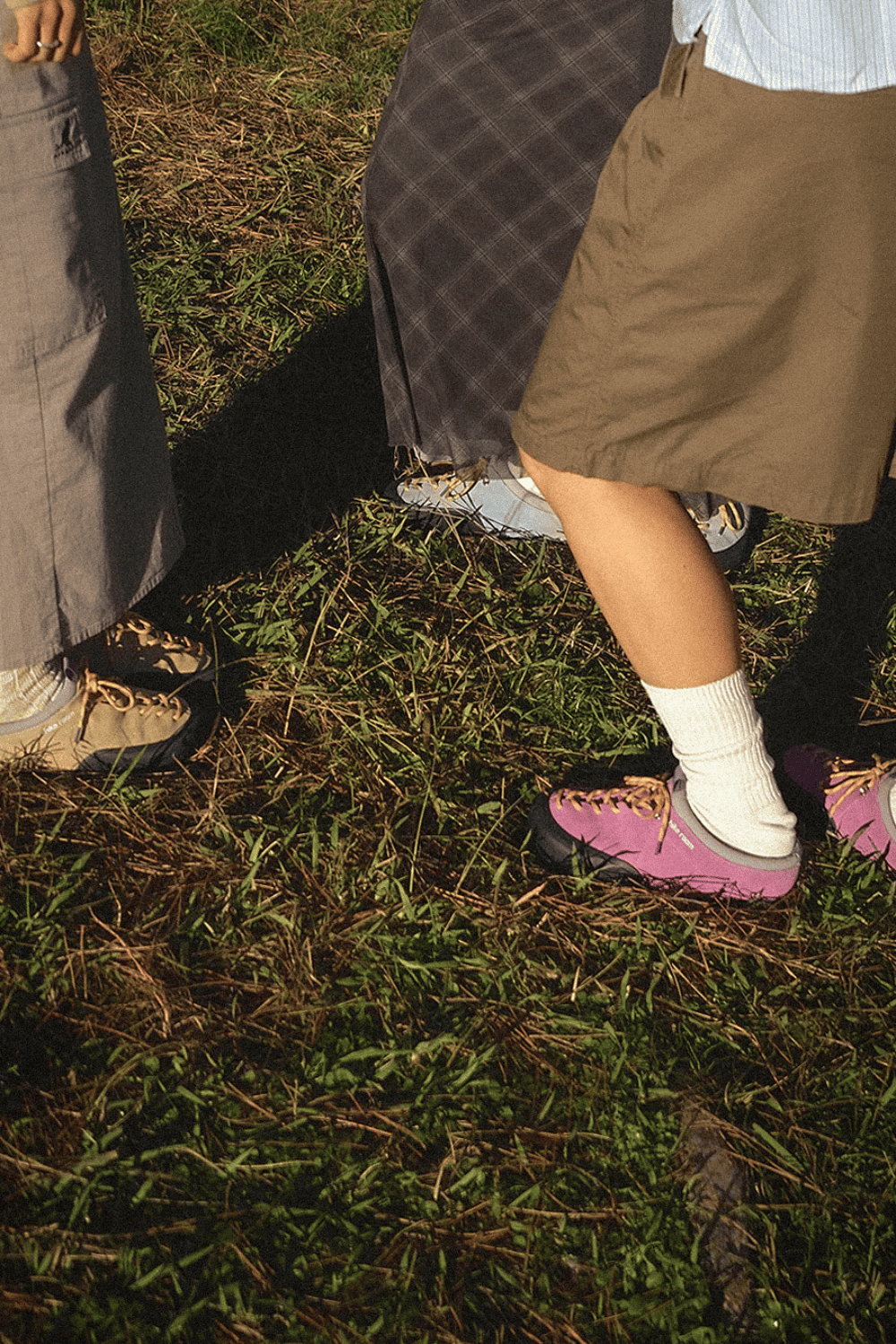 Piernas de tres personas de pie sobre la hierba vistiendo faldas y pantalones cargo combinados con diferentes zapatillas Hike Room en tonos beige, gris y morado.