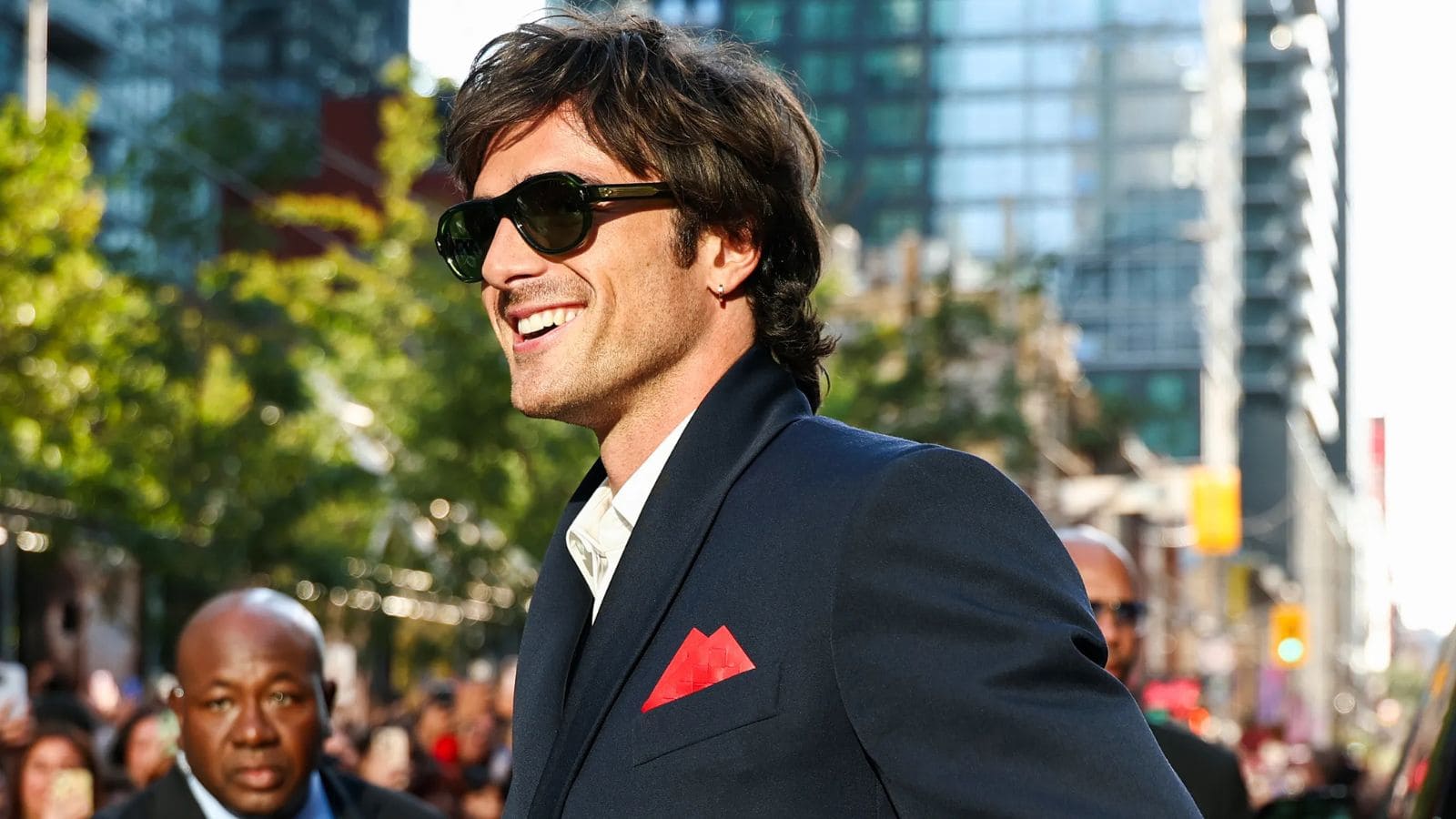 Jacob Elordi luciendo gafas de sol negras con traje oscuro y pañuelo rojo durante un evento en la ciudad