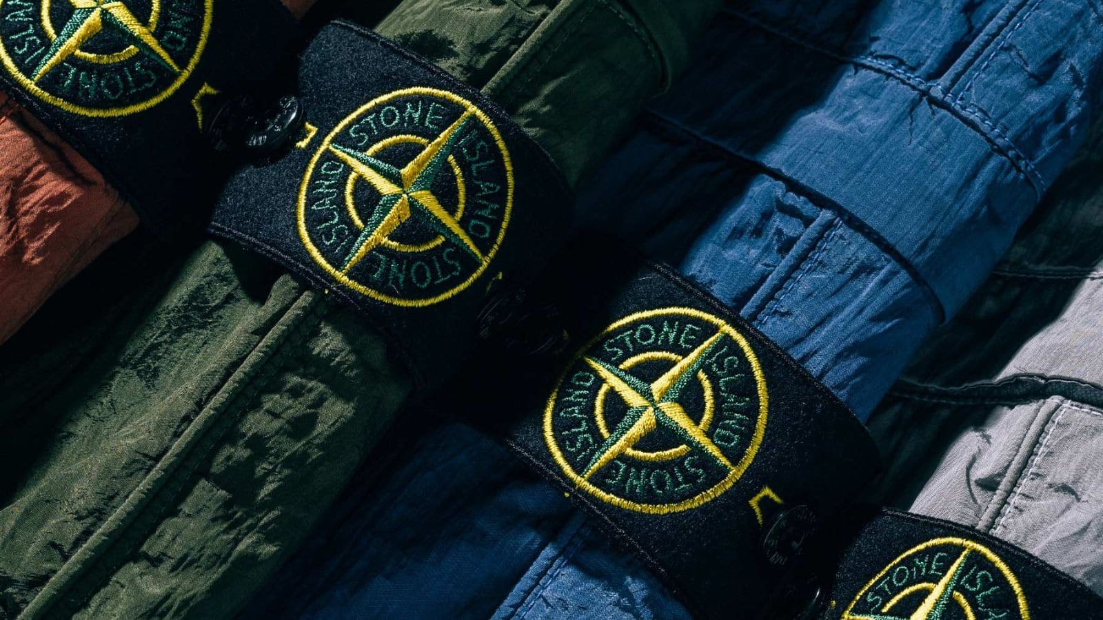 Primer plano del parche con brújula de Stone Island sobre chaquetas técnicas en distintos colores.