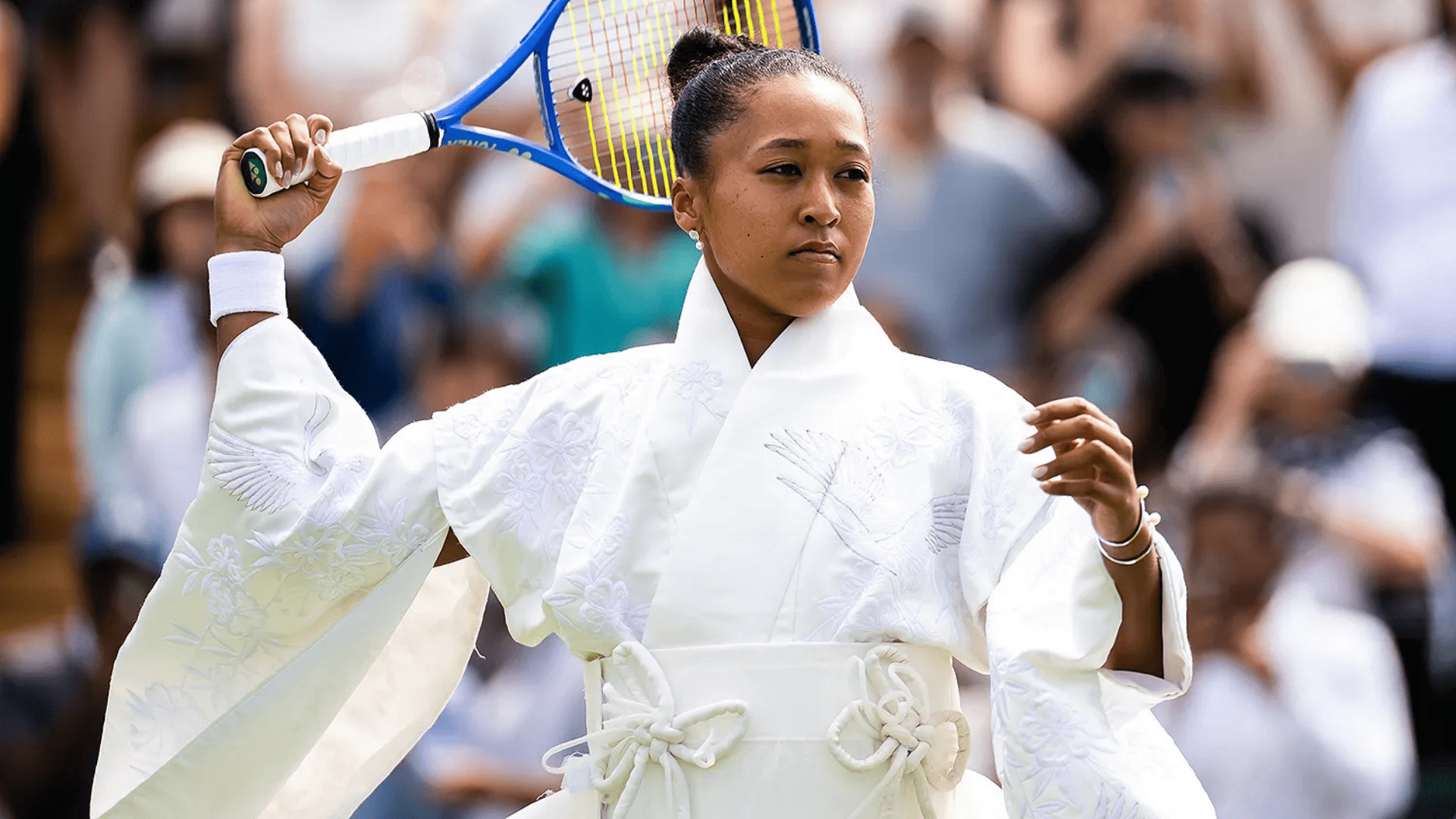 Primer plano de la tenista Naomi Osaka en la pista preparándose para sacar, vistiendo un abrigo estilo kimono blanco con bordados