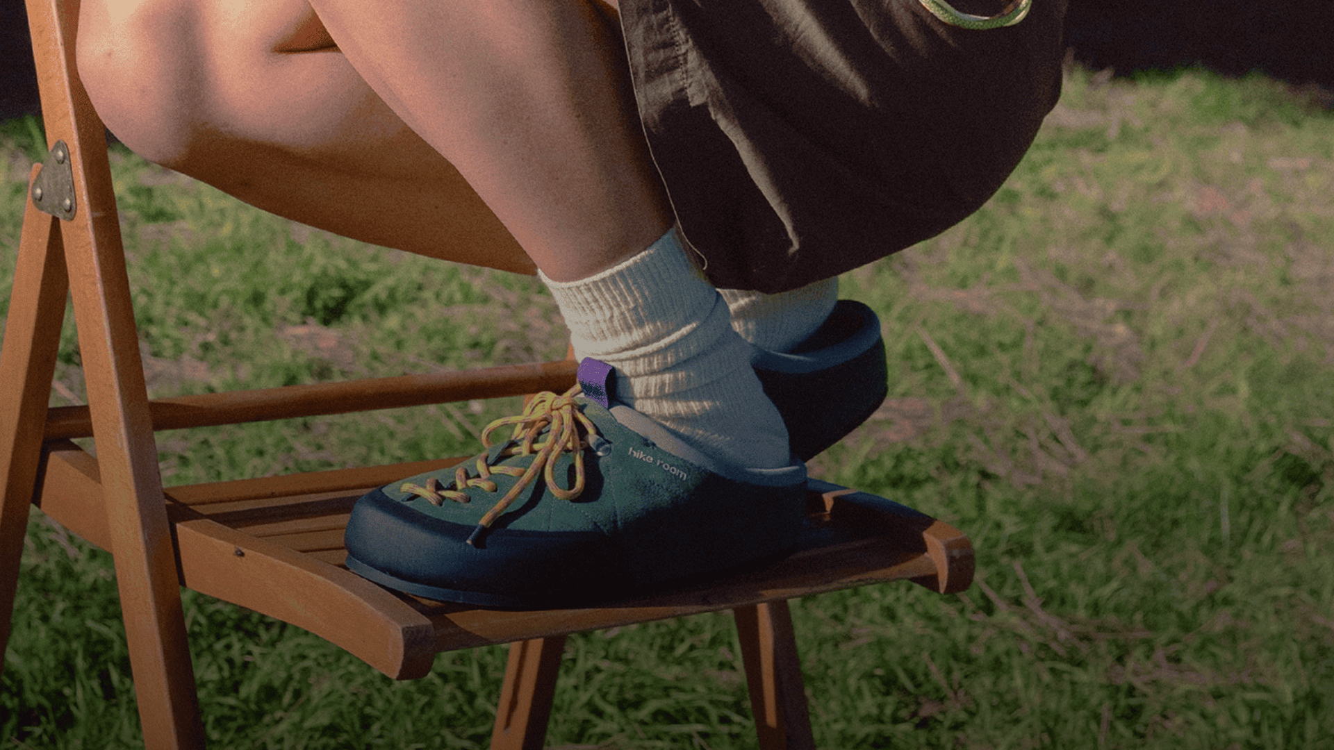 Persona sentada en una silla de madera de camping en la naturaleza calzando las zapatillas tipo zueco verdes Extramonta Pine Green de Hike Room con calcetines blancos.