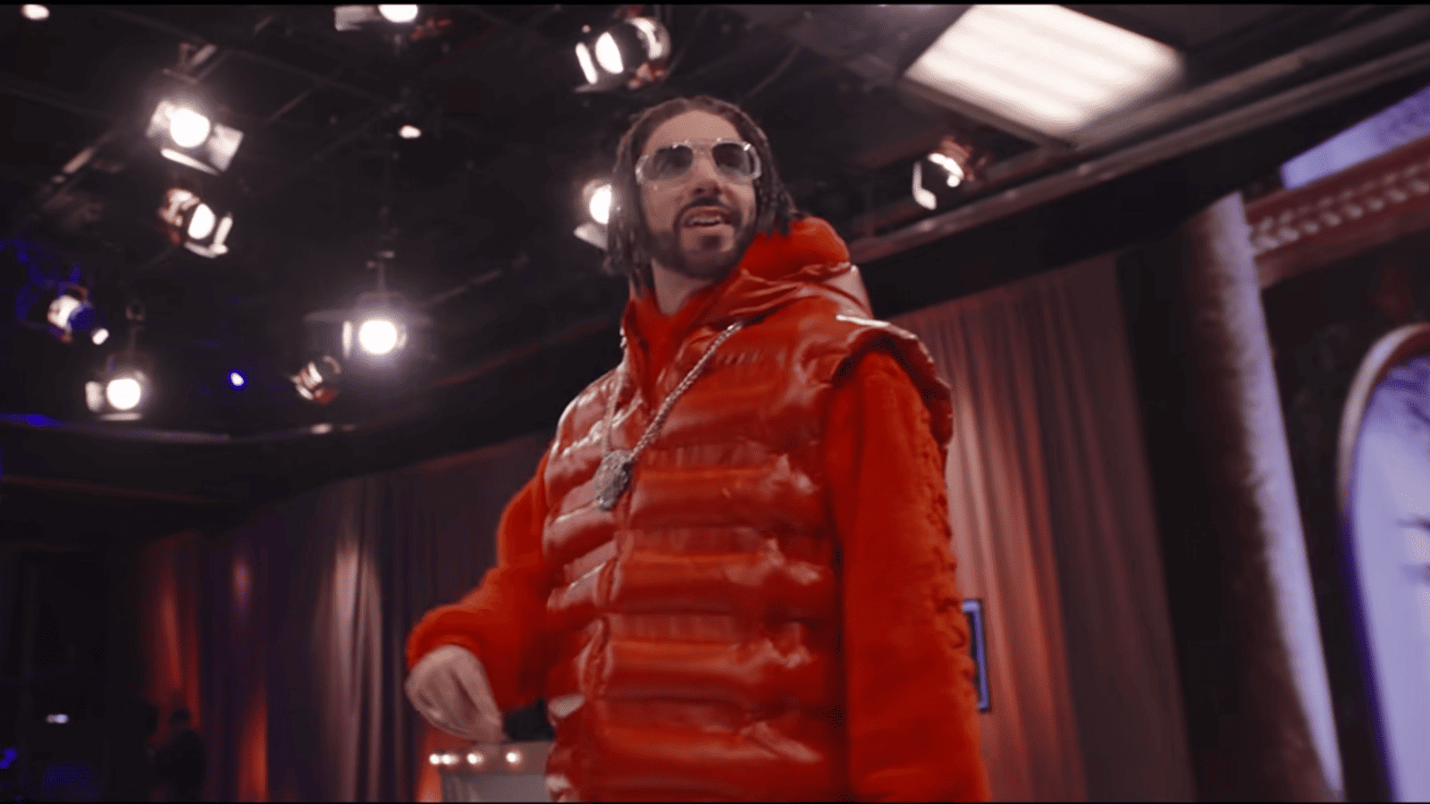 Ali G con chaleco acolchado rojo brillante, jersey rojo y gafas transparentes oversize en un plató de Ali G: Who Iz I