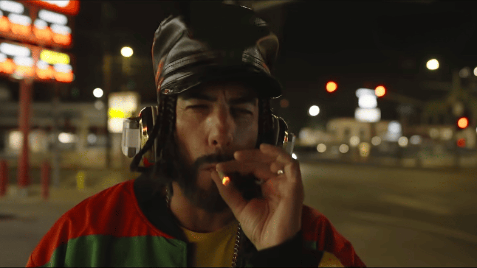 Ali G con bomber en colores rasta, gorra de cuero negra y cascos plateados fumando en la calle de noche en Ali G: Who Iz I